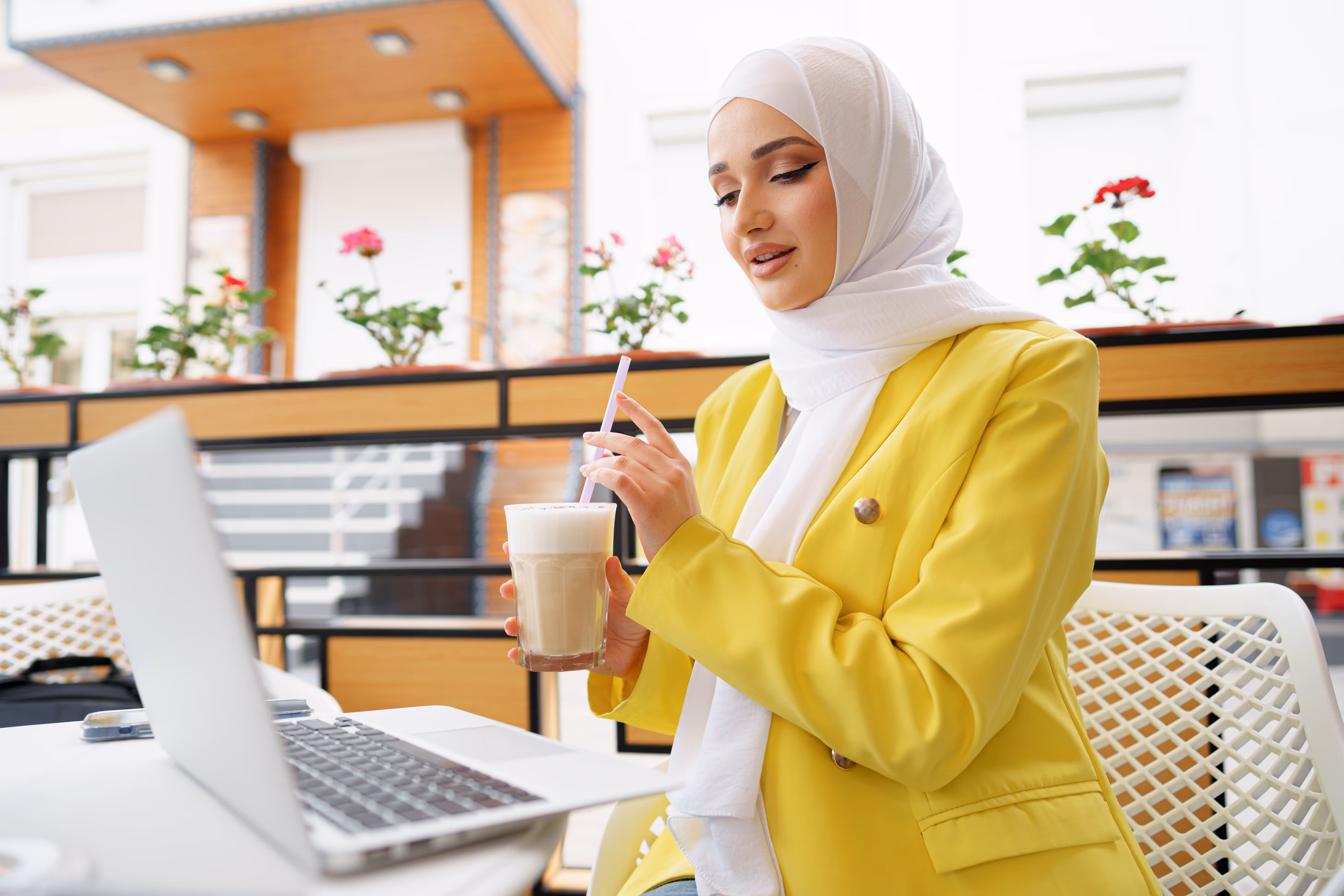 Young woman wearing a white hijab and yellow blazer sitting at a cafe table with a laptop, holding a glass of iced coffee with a straw.