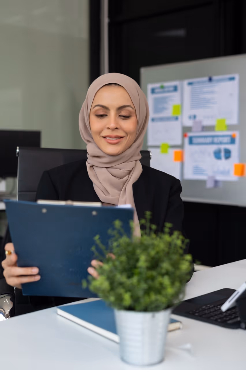 Smiling businesswoman wearing a hijab seated at a desk reviewing a clipboard, with a laptop and green plant in front and a board with reports in the background.