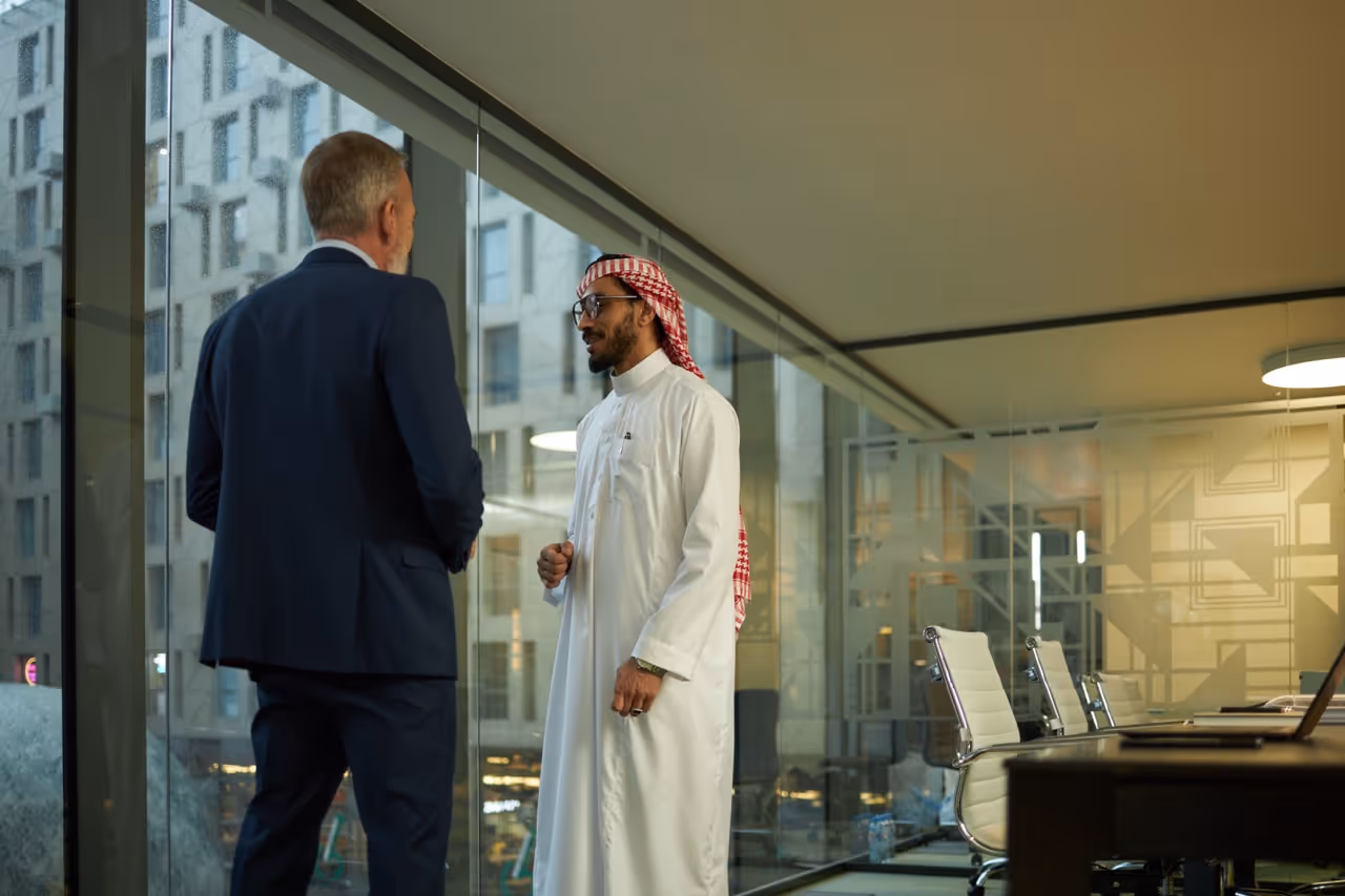 Two businessmen, one in a navy suit and the other in traditional Middle Eastern attire, talking in a modern office with glass walls.