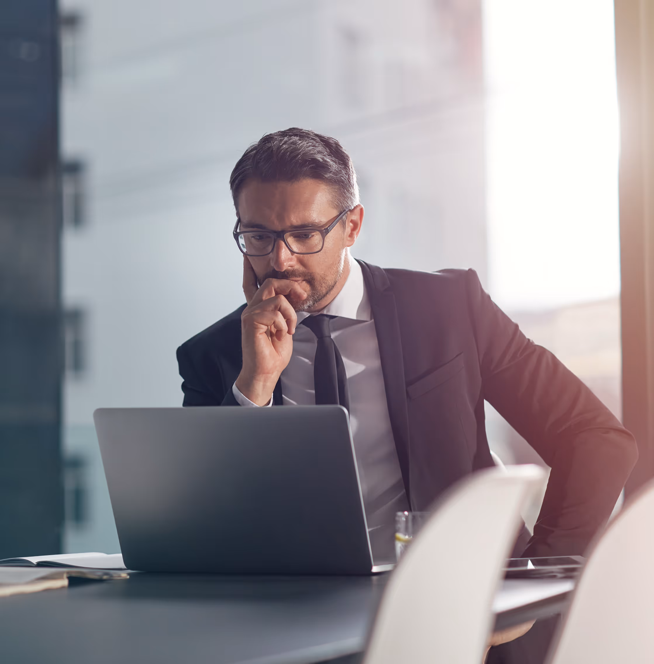 Businessman in glasses and suit thoughtfully looking at laptop screen in bright office.
