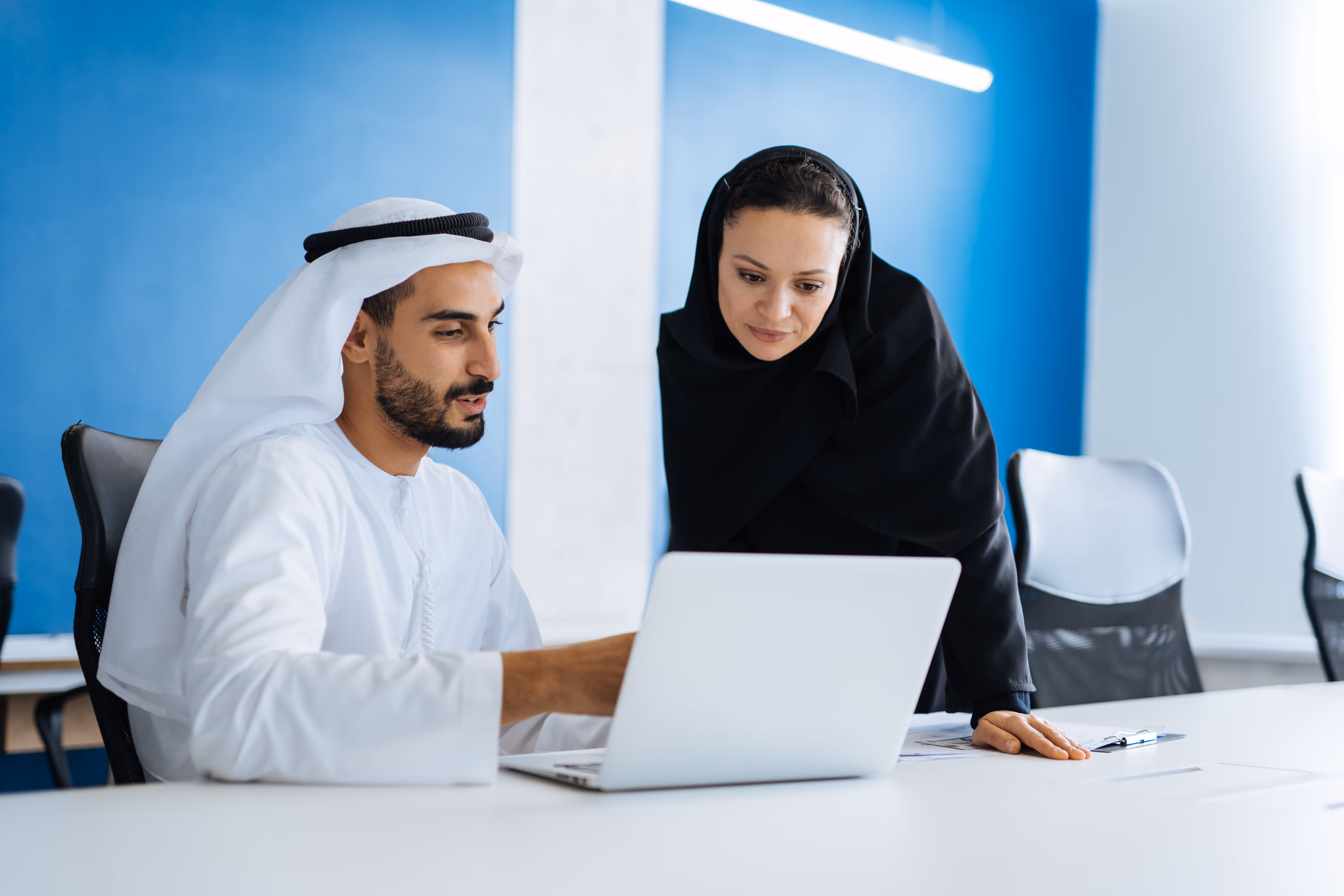 Man in traditional Middle Eastern attire and woman in black hijab reviewing something on a laptop in a modern office.