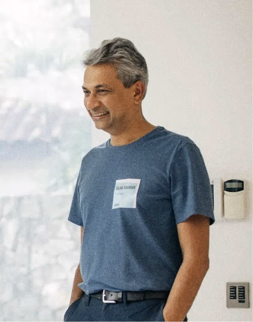 Smiling man with gray hair wearing a blue t-shirt and black belt standing indoors near a white wall.