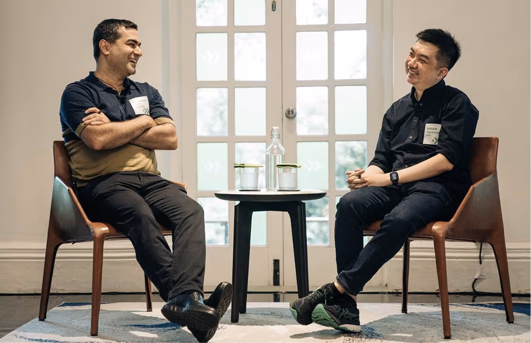 Two men sitting on chairs facing each other and smiling with a small table holding a bottle and two glasses between them.