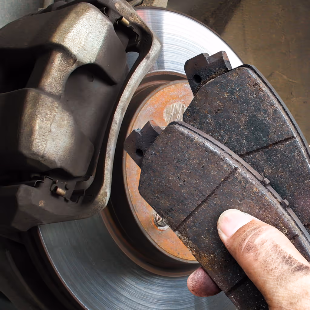 A hand holding two heavily worn brake pads next to a brake disc, illustrating the need for replacement during a Maxamy Mobile Autocare service.