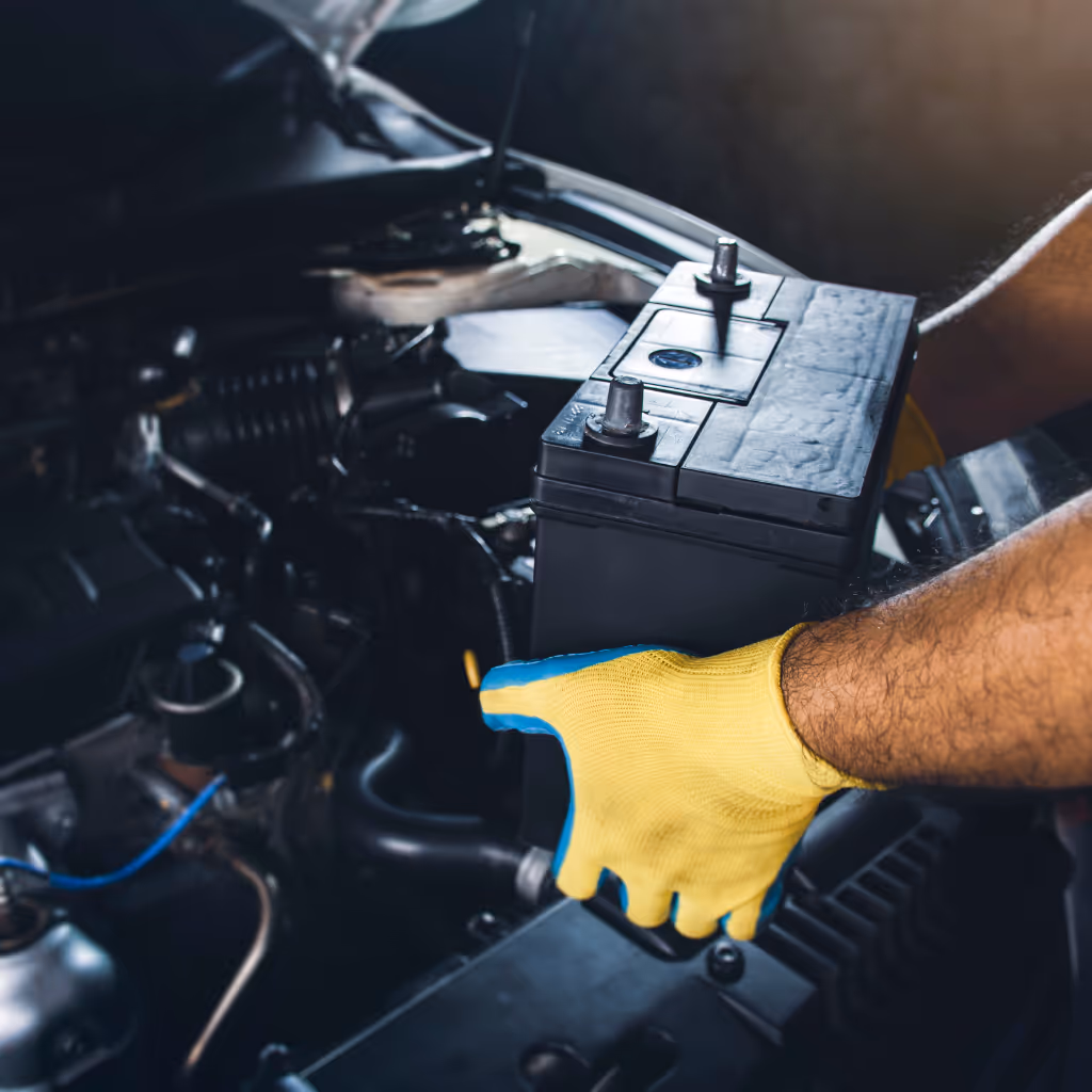 A Maxamy Mobile Autocare professional wearing yellow gloves is working on the electrical connections of a car's headlight assembly.