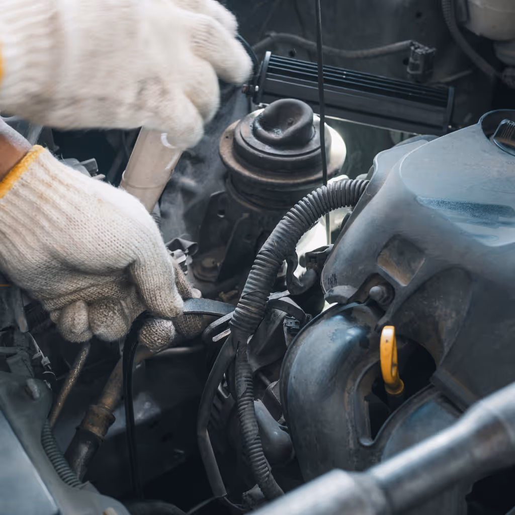 Gloved hands of a Maxamy Mobile Autocare technician carefully inspect an engine drive belt for wear, cracks, and proper tension during a routine check.