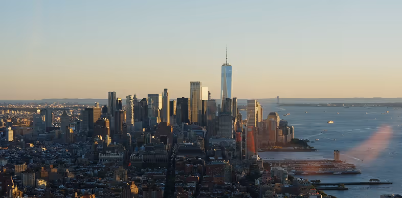 Panoramic view of Lower Manhattan skyline at sunset, including One World Trade Center and surrounding buildings by the waterfront.