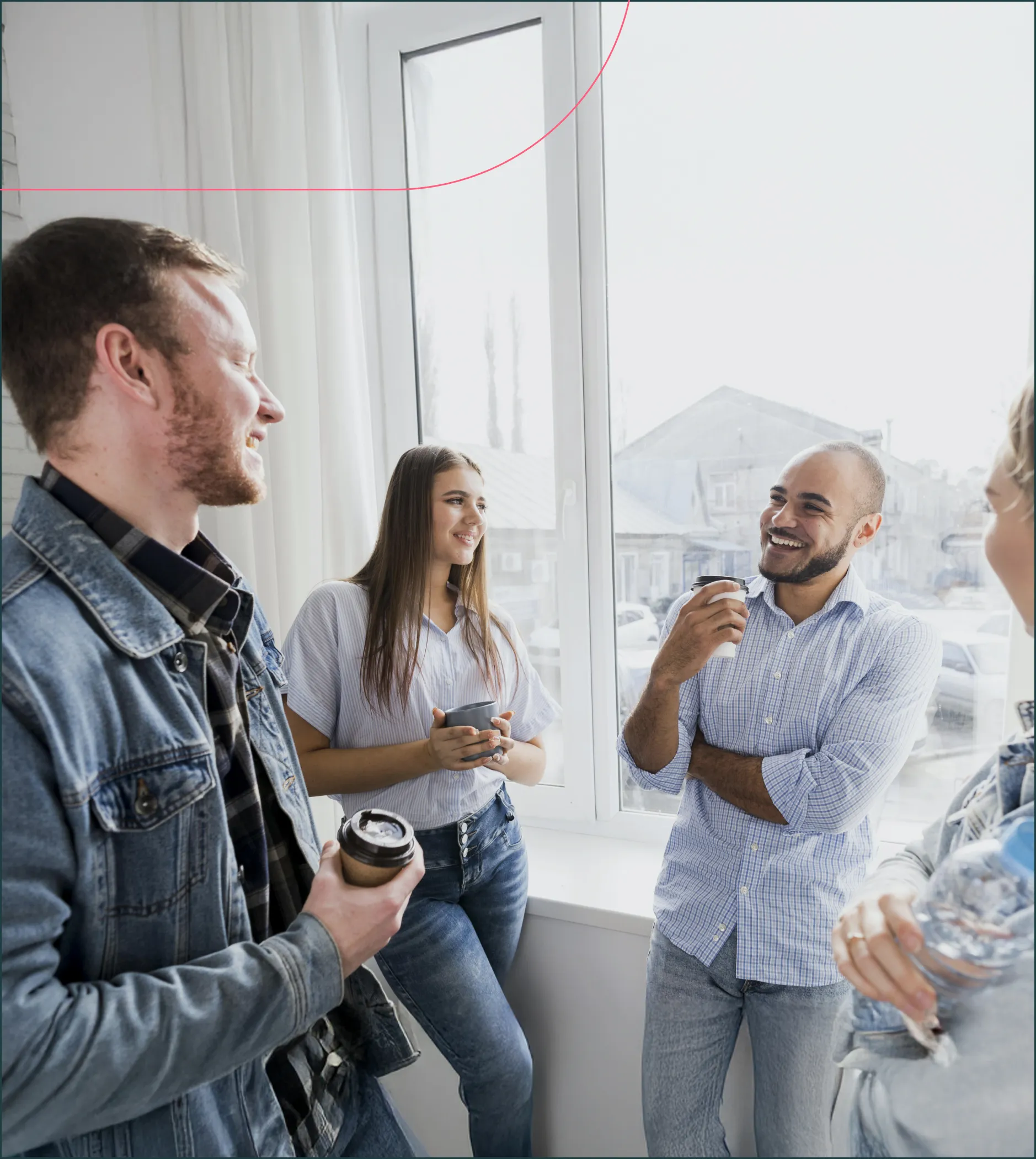 Four casually dressed young adults laughing and chatting while holding drinks near a large window.