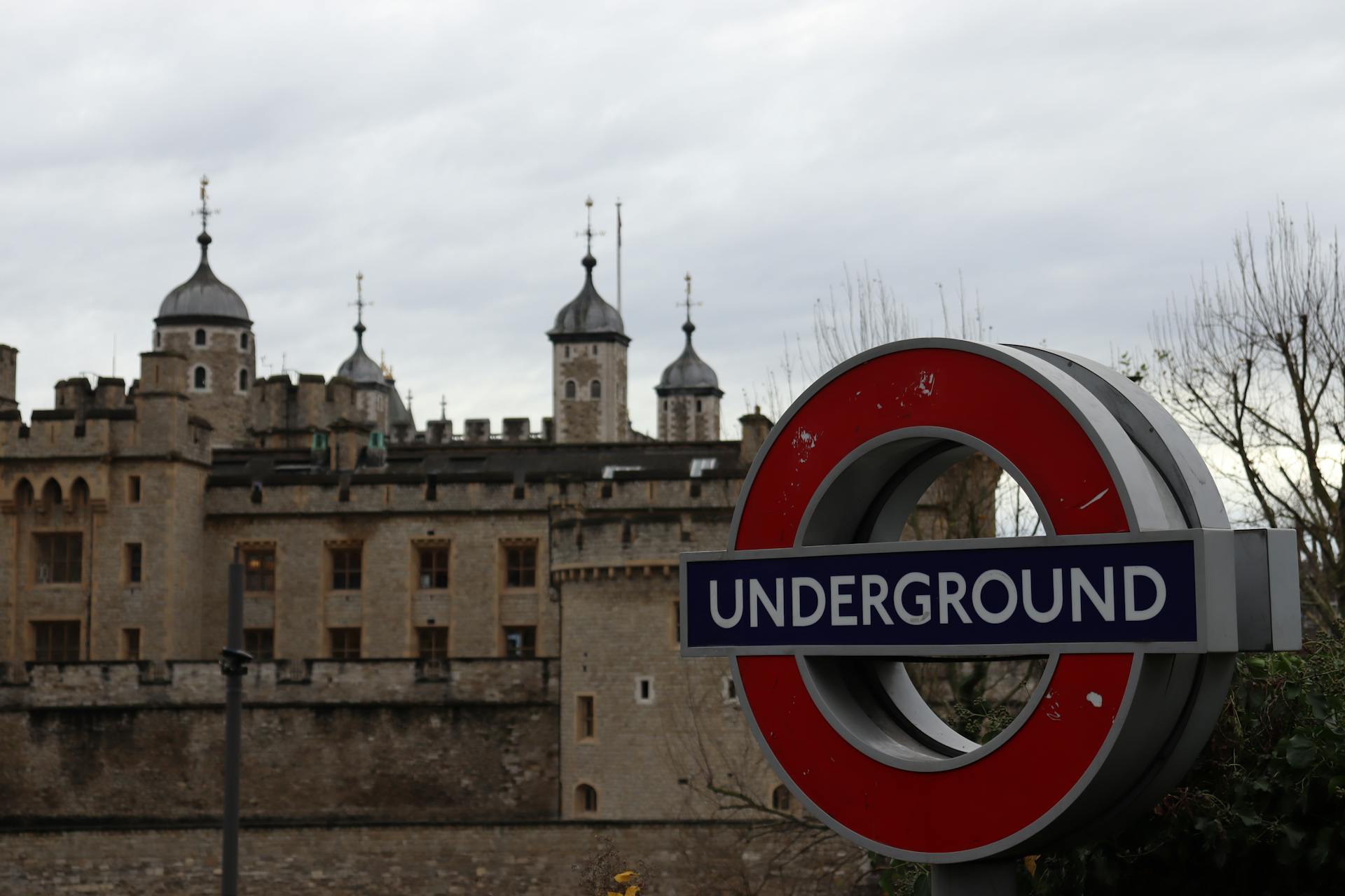 Red London Underground sign in front of the historic Tower of London under a cloudy sky.