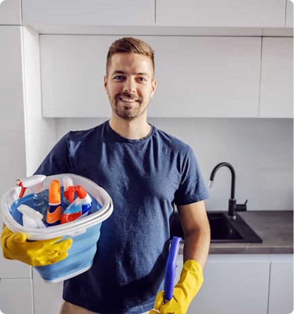 Smiling man in a navy shirt holding a blue bucket with cleaning supplies and wearing yellow gloves in a modern kitchen.