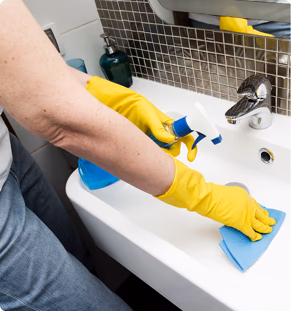 Person wearing yellow rubber gloves cleaning a white bathroom sink with a blue cloth and spray bottle.