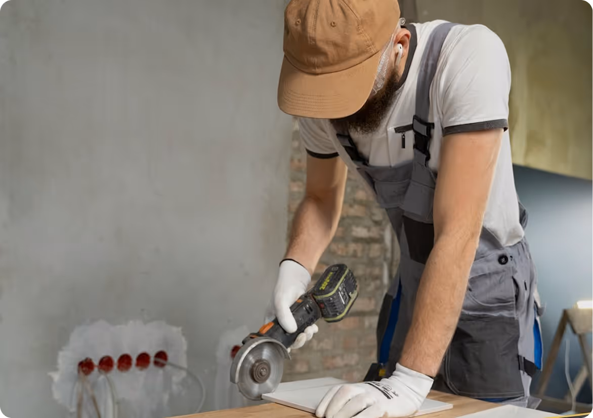 Construction worker wearing a brown cap, gloves, and overalls using a power saw to cut a tile indoors.