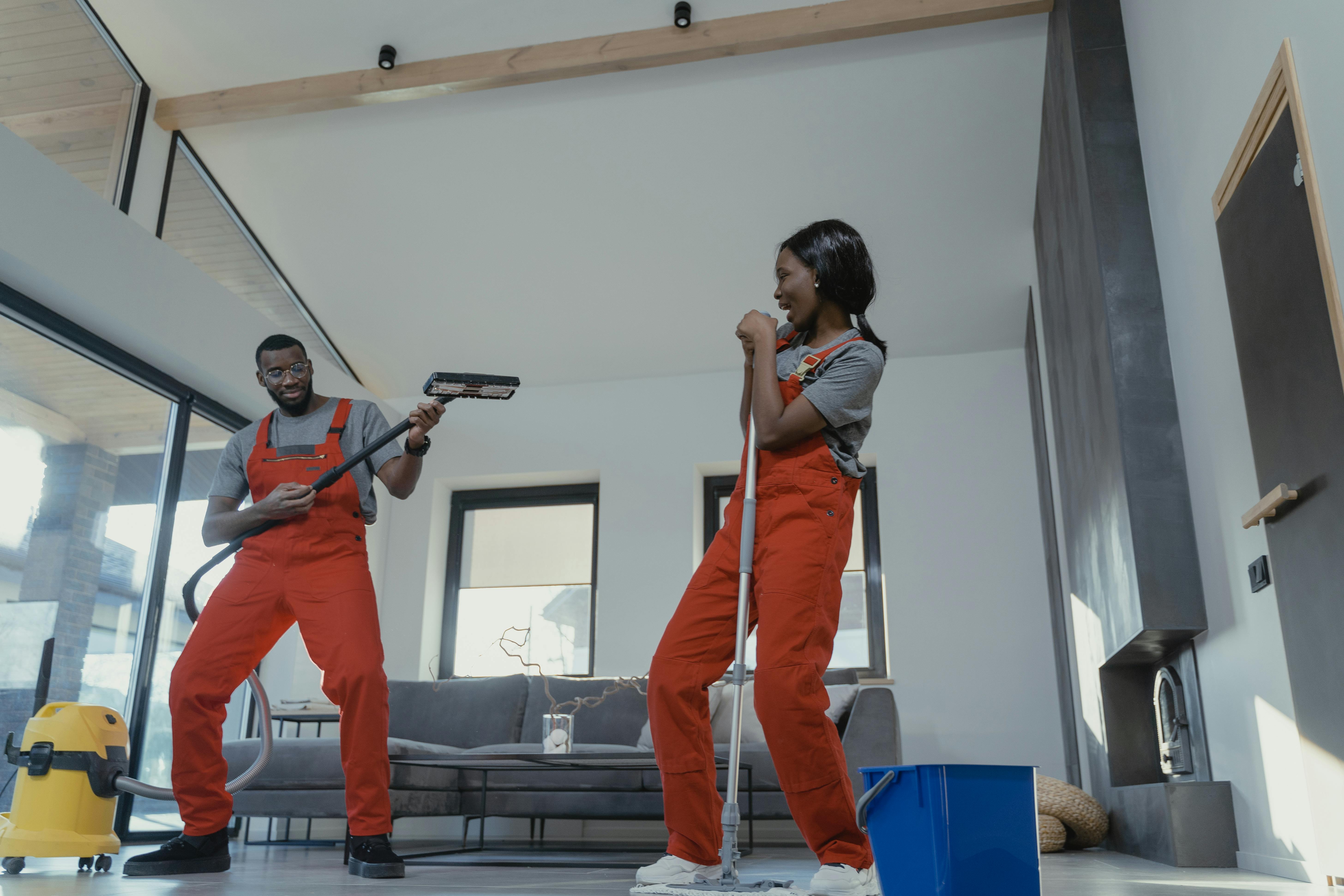 Two people in red overalls joyfully cleaning a modern living room, one holding a vacuum cleaner hose like a guitar and the other holding a mop.