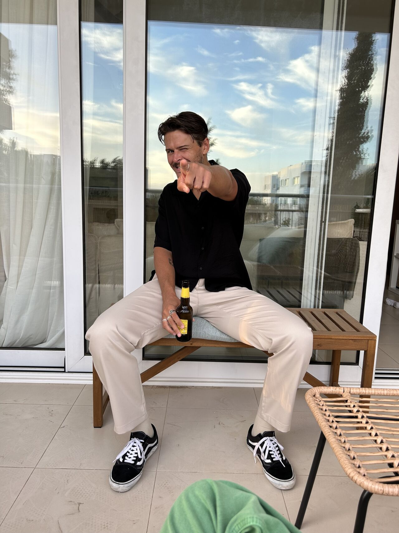 Man sitting on a wooden bench pointing forward with a beer bottle in one hand, wearing a black shirt, beige pants, and black sneakers.