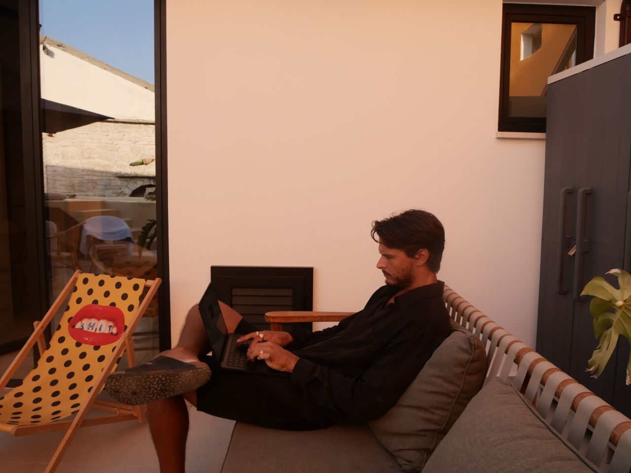 Man in black shirt and shorts sitting on a patio sofa working on a laptop with one leg resting on a polka-dotted deck chair.