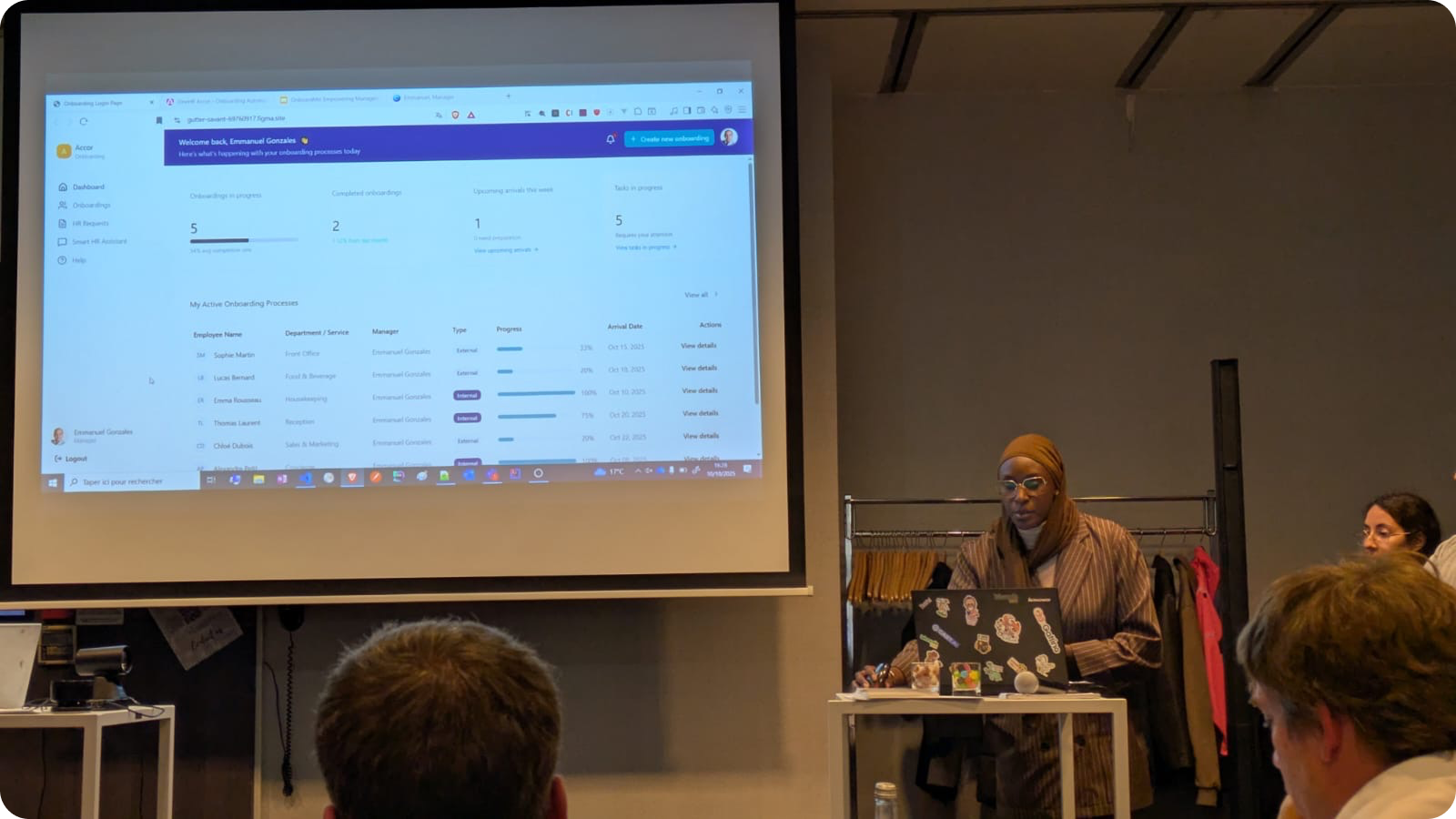 A woman wearing a headscarf presents onboarding progress data displayed on a projector screen to an audience.