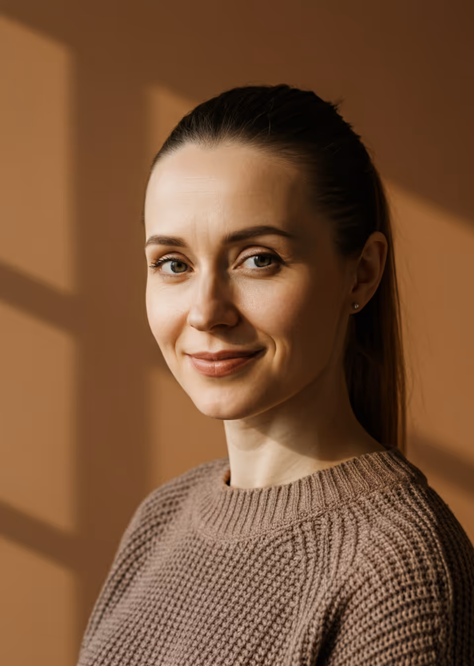 Smiling woman with light skin and light brown hair tied back, wearing a brown knit sweater against a brown background with window shadows.