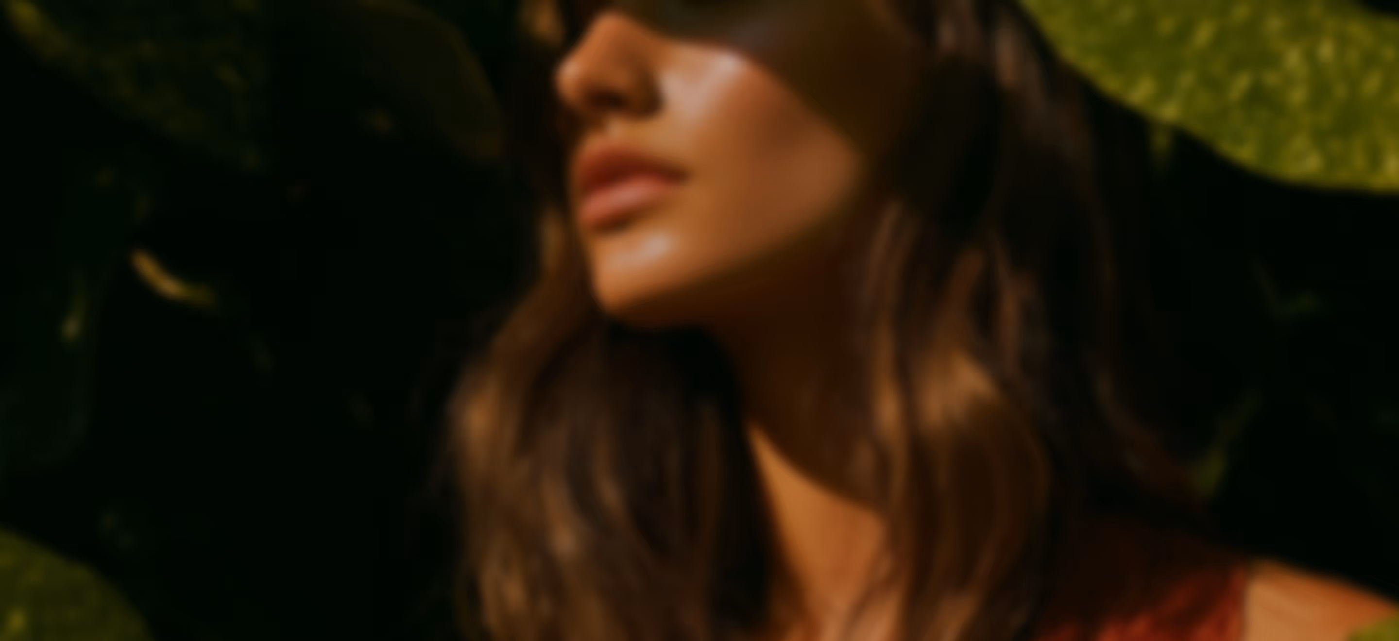 Close-up of a woman's face and hair with shadow patterns and green leaves in the background.