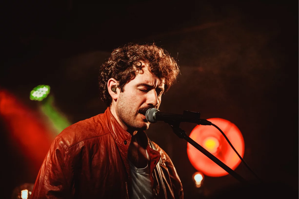 Ein Mann mit Locken und Schnurrbart singt mit geschlossenen Augen in ein Mikrofon bei einem Konzert.