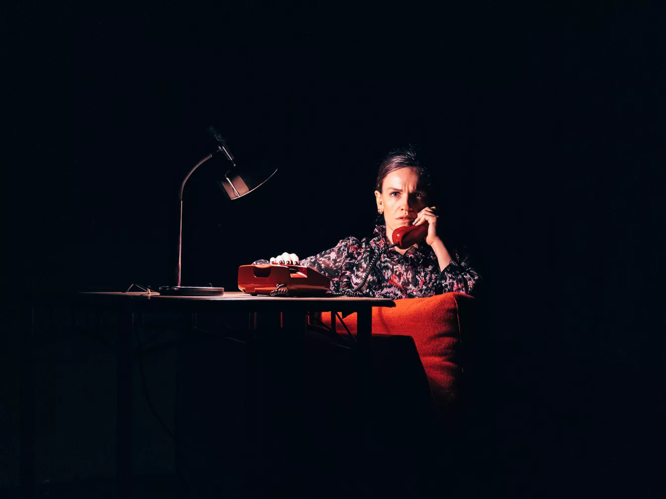 Frau mit besorgtem Gesichtsausdruck spricht an einem roten Telefon in dunklem Raum mit Schreibtischlampe.