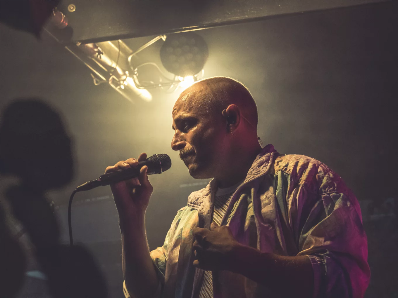 Sänger mit Glatze singt leidenschaftlich in ein Mikrofon auf einer dunklen Bühne mit Bühnenbeleuchtung.