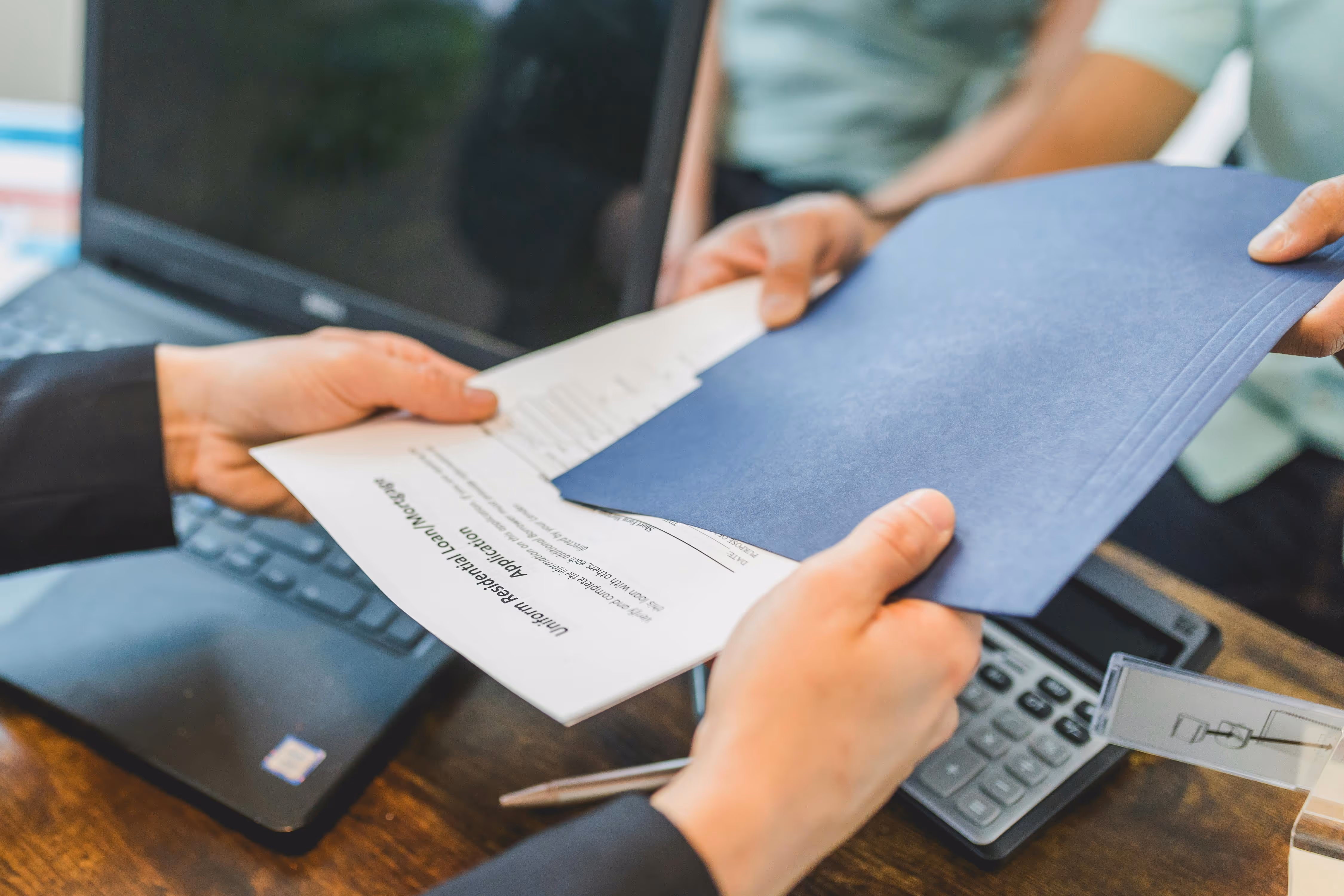 Hands exchanging a blue folder and documents on a desk with a laptop and calculator in the background.
