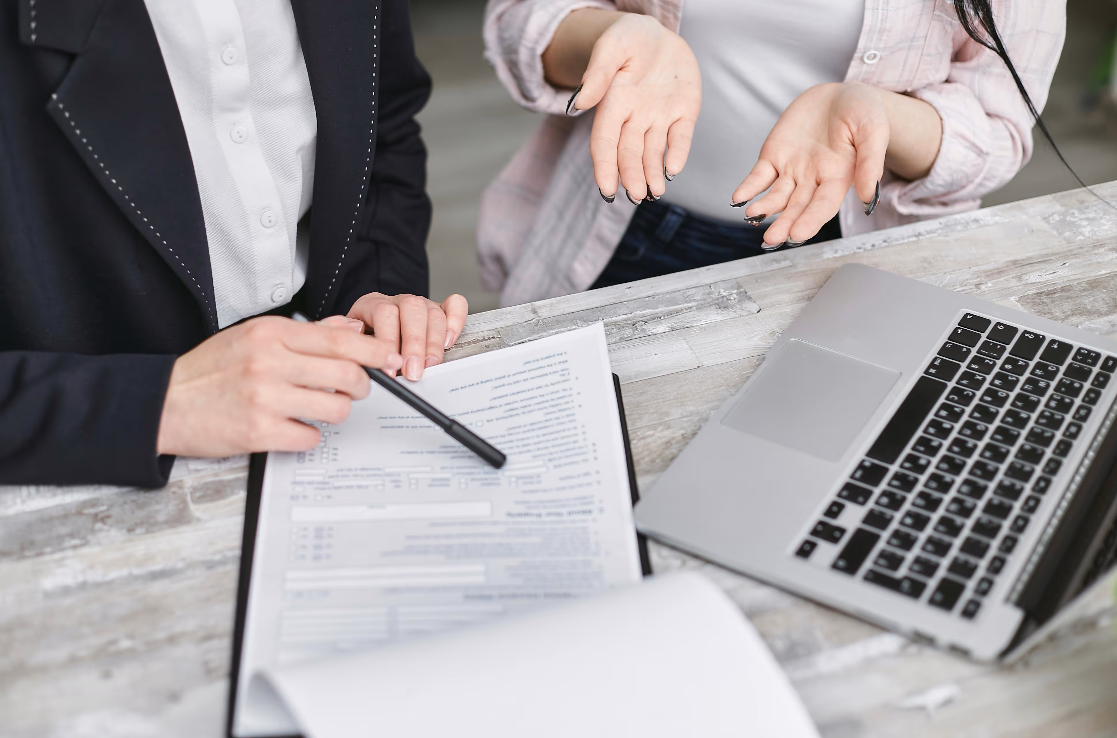 Two people at a desk reviewing a document on a clipboard next to a laptop, one person pointing with a pen and the other gesturing with open hands.
