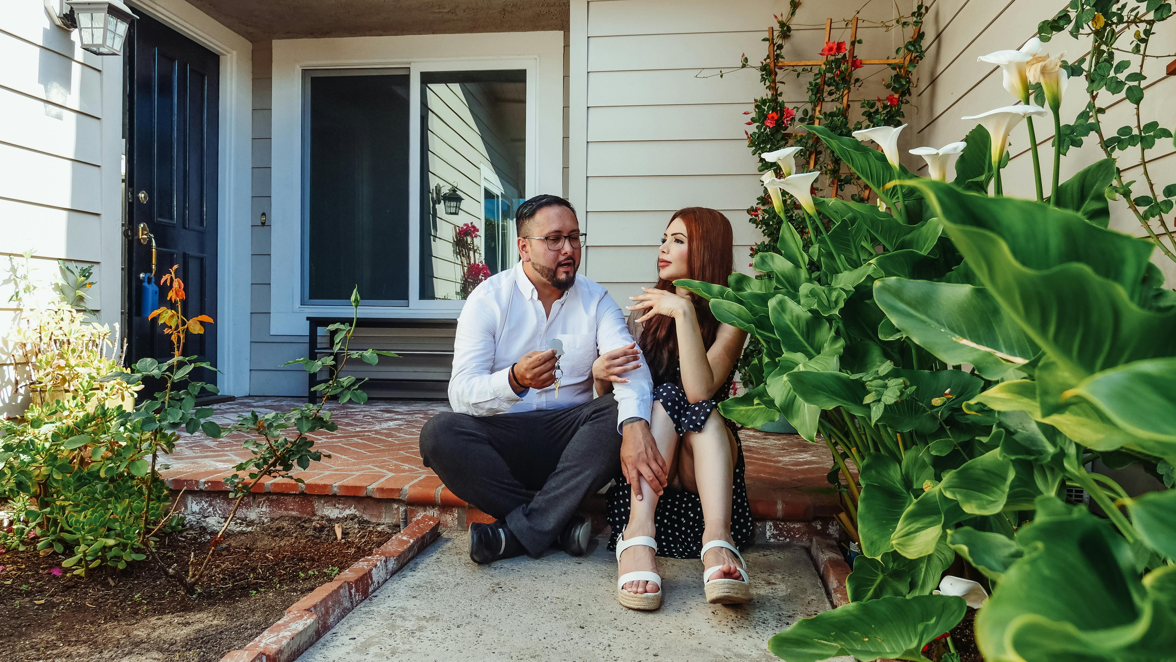 Man holding house keys sits on porch next to woman with red hair, surrounded by plants and flowers.