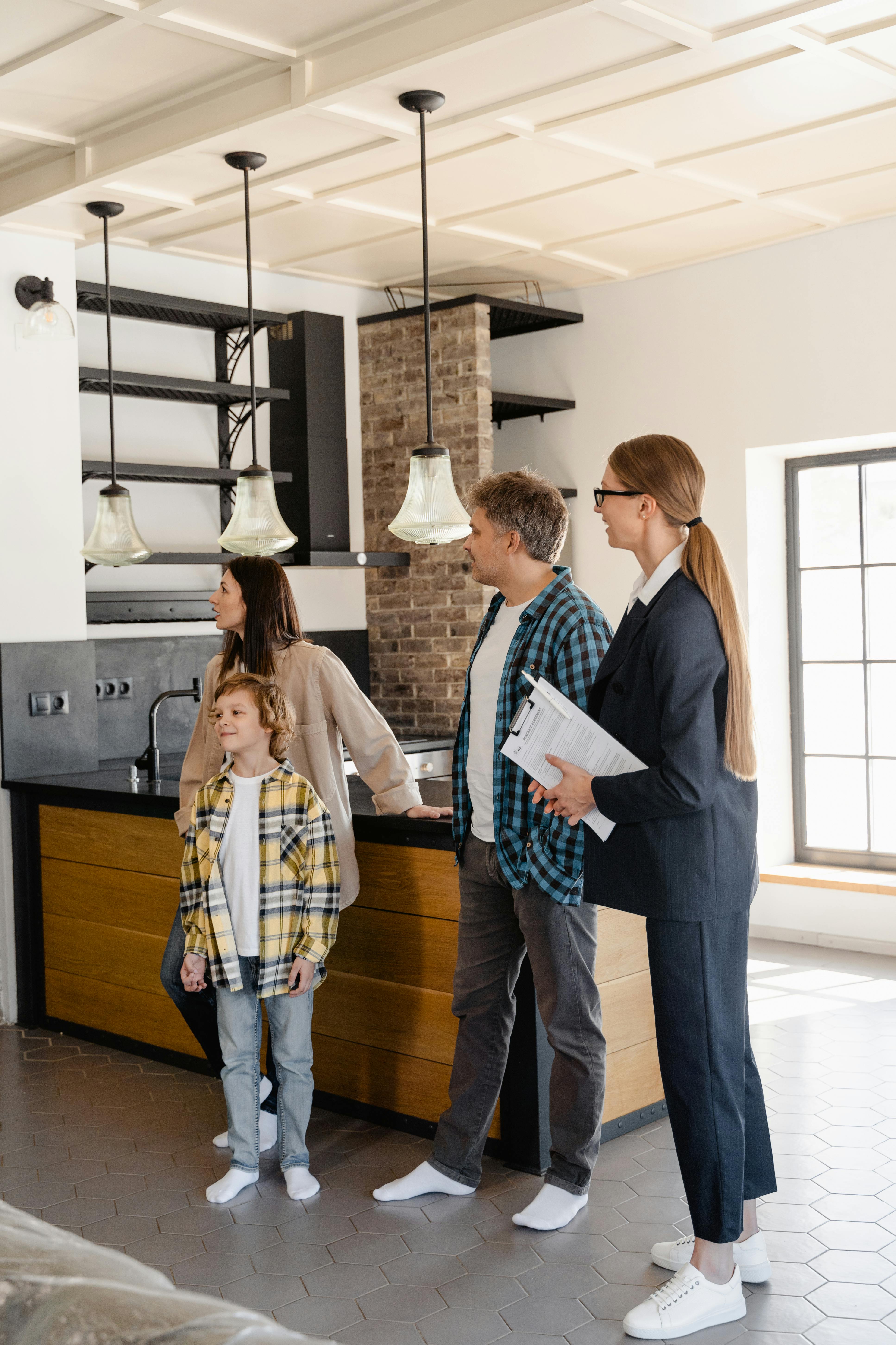 A family with a young boy looks around a modern kitchen while a woman in business attire holds a clipboard, presumably a real estate agent.