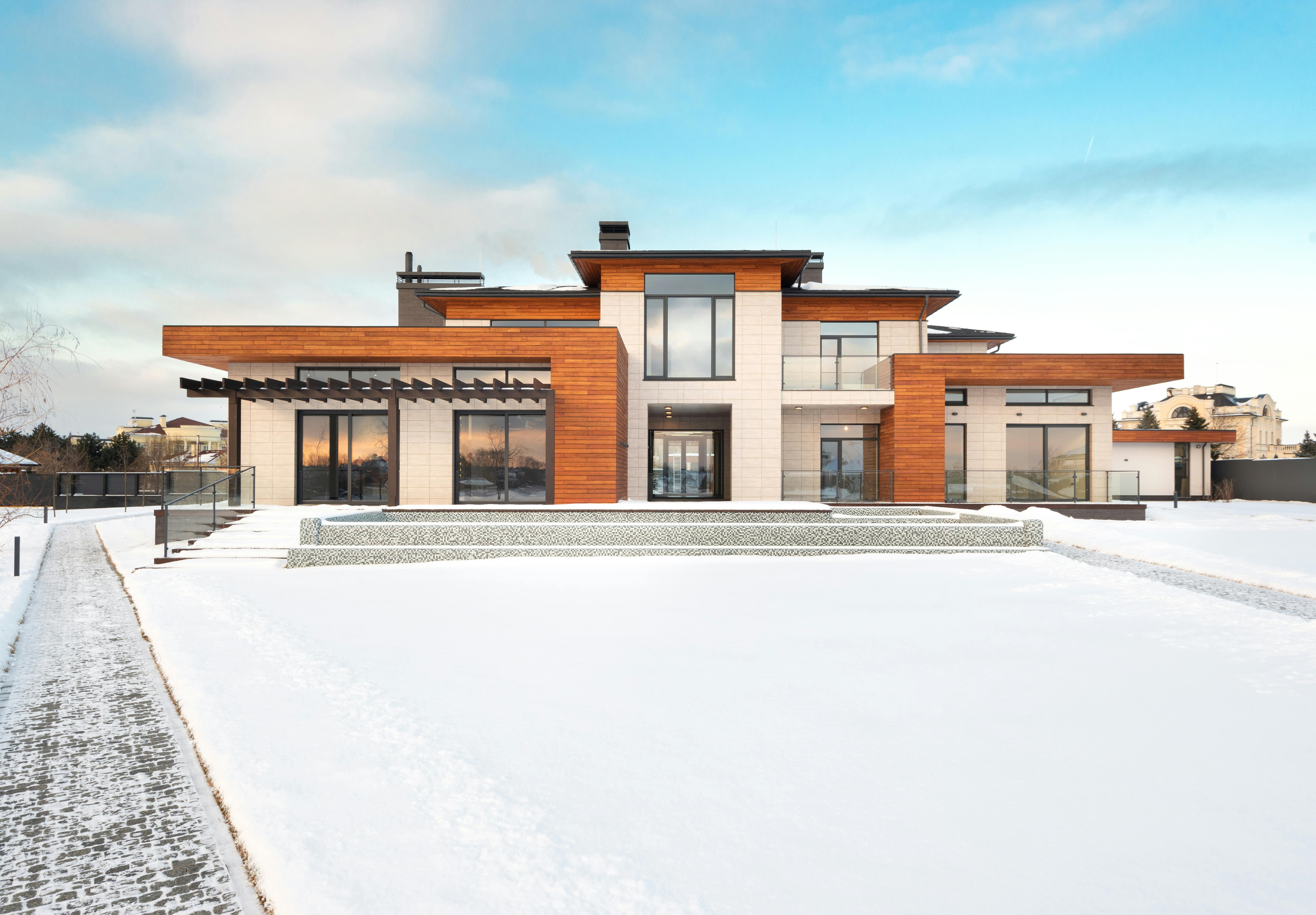 Modern two-story house with large windows, wood and stone exterior, surrounded by snow and a paved walkway.