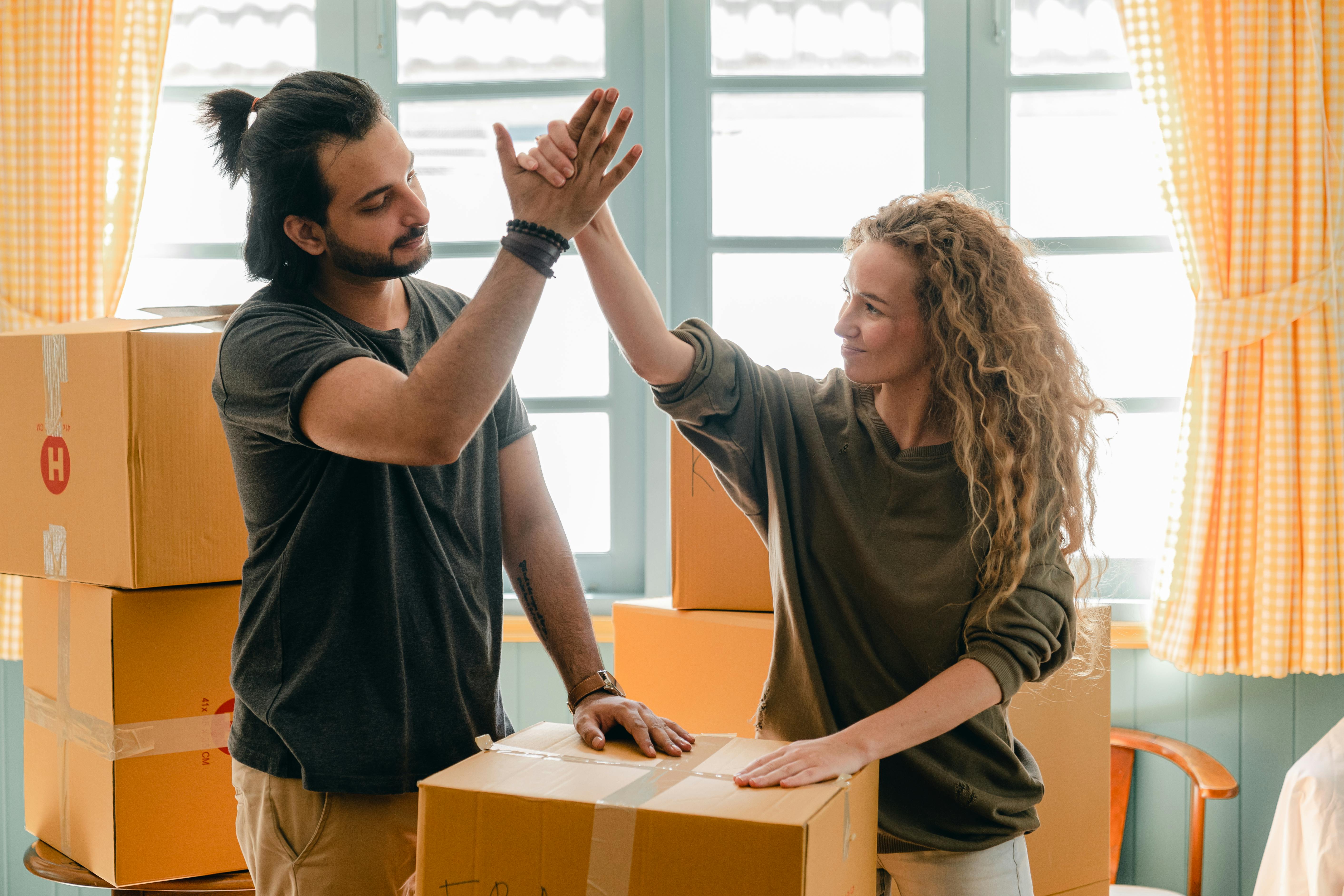 Young man and woman giving a high-five while standing among cardboard moving boxes in a sunlit room.