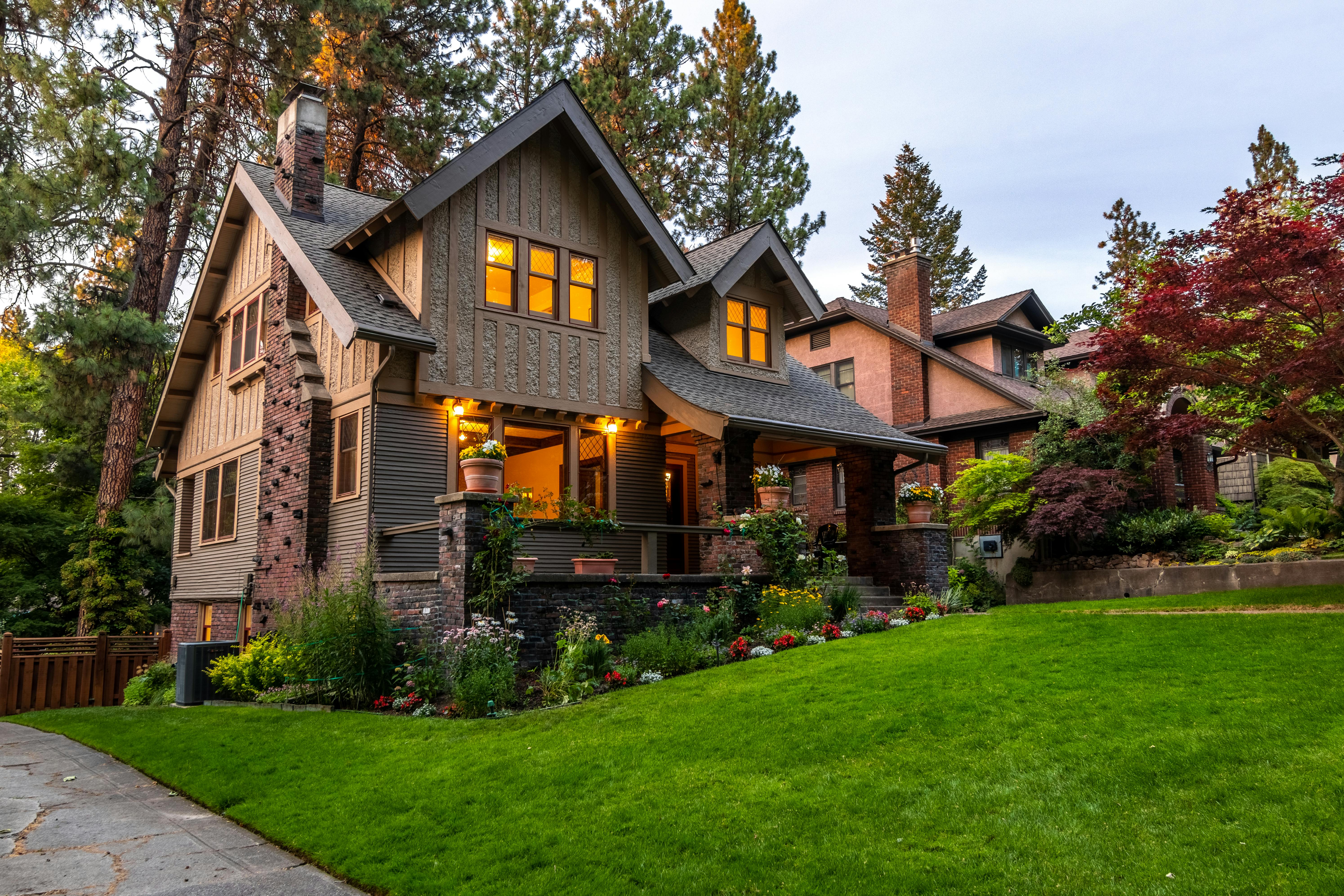 Two-story craftsman-style house with lit windows, a well-kept green lawn, and colorful flower beds in the front yard.