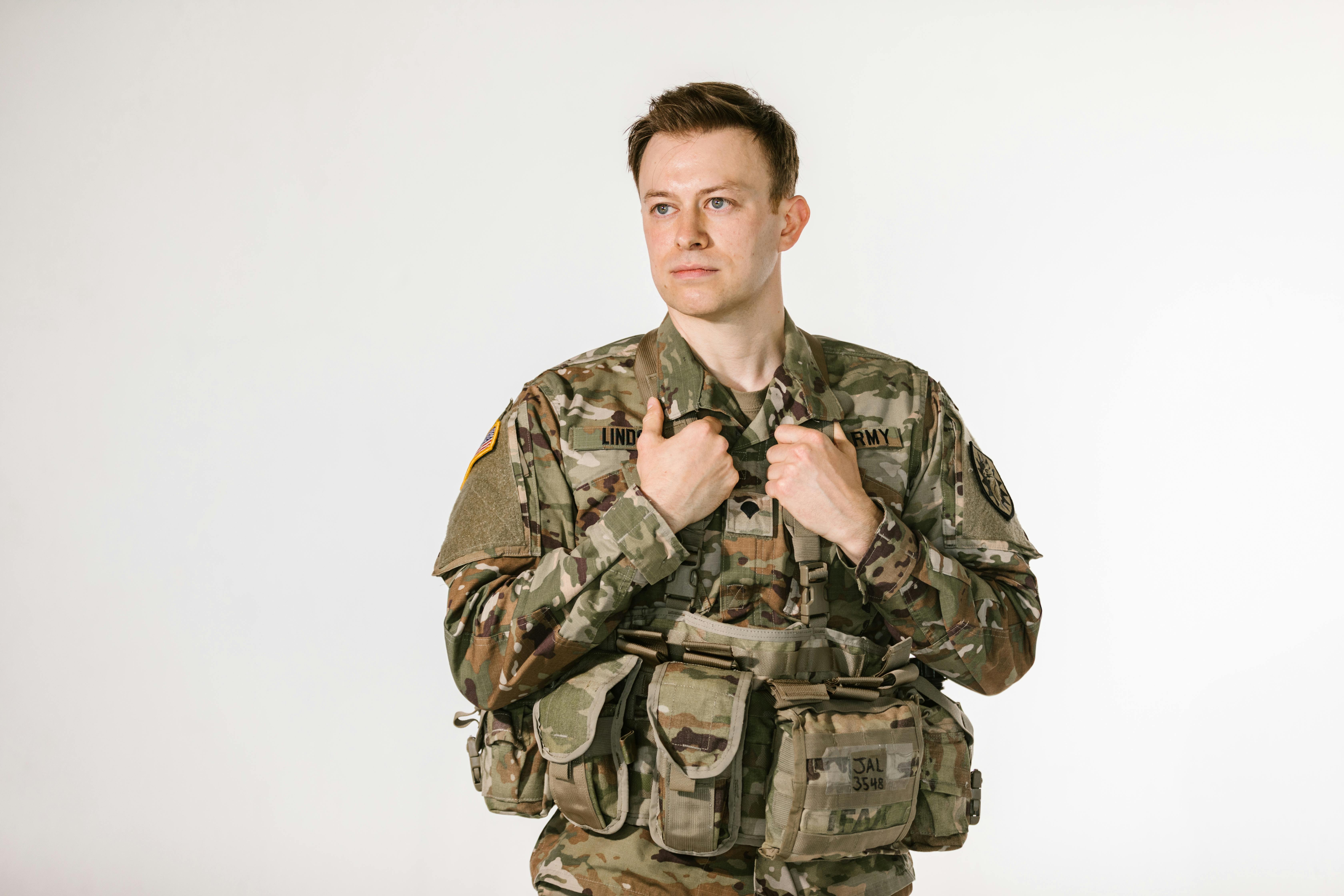 A man in camouflaged military uniform holding shoulder straps against a plain white background.