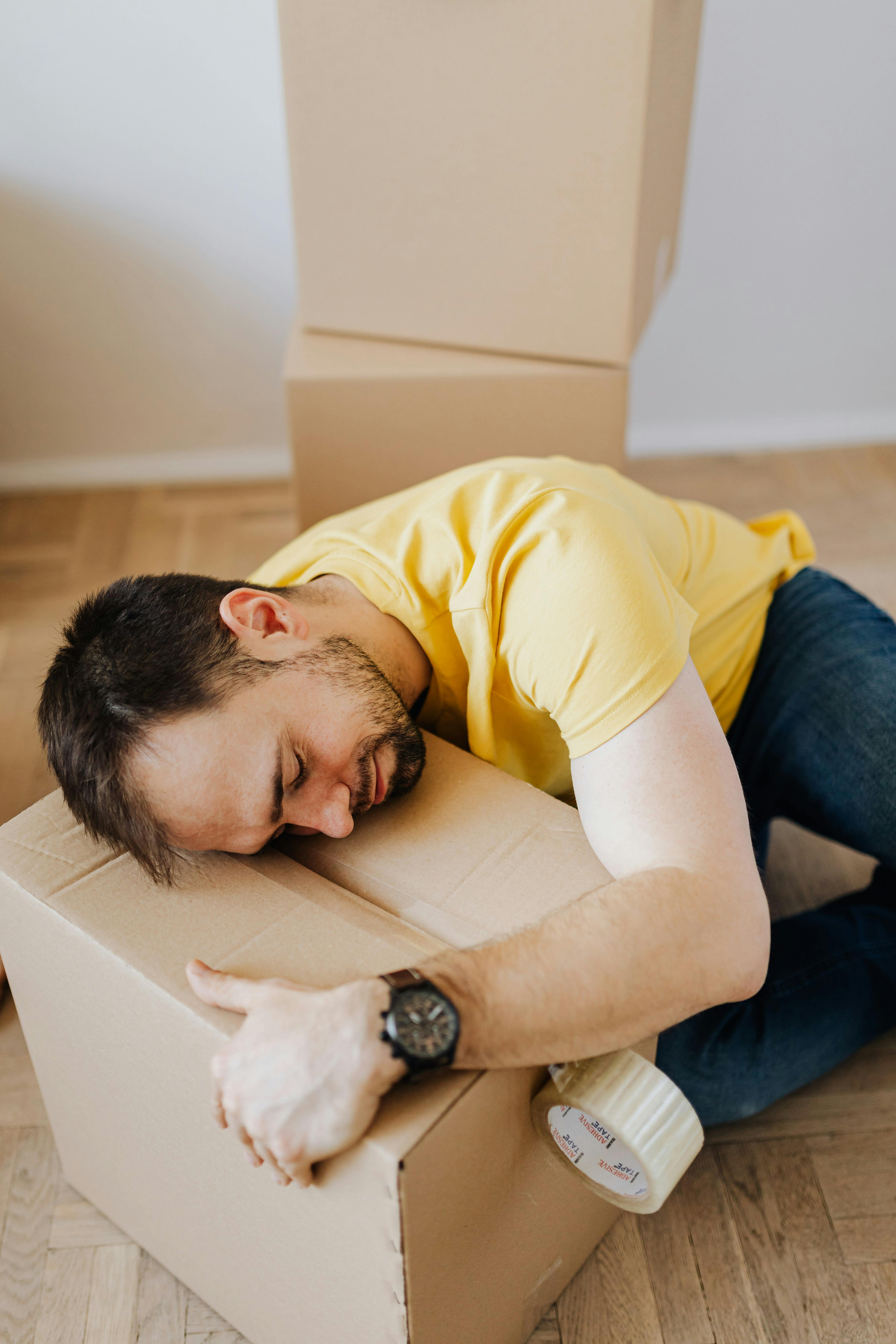 Man in a yellow shirt hugging a cardboard box while sitting on the wooden floor with packing tape nearby.