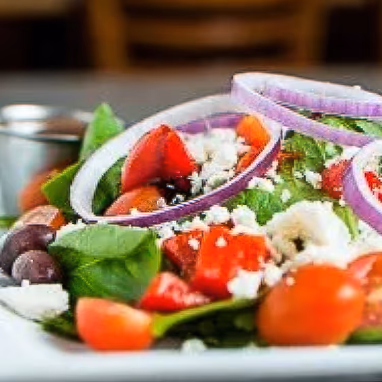 Close-up of a fresh salad with spinach, cherry tomatoes, red onion rings, feta cheese crumbles, and Kalamata olives on a white plate.