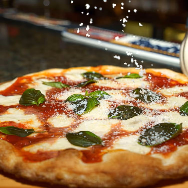 Margherita pizza topped with fresh basil leaves and grated cheese being sprinkled on top.