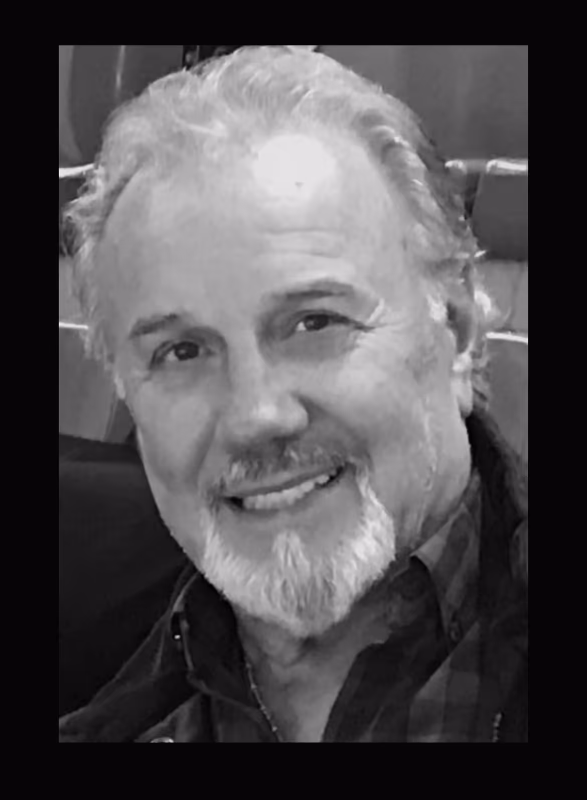 Smiling older man with white hair and beard wearing a dark checkered shirt.