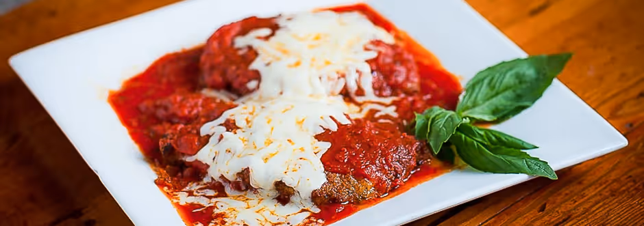 Two pieces of meat topped with melted mozzarella cheese and marinara sauce, garnished with fresh basil leaves on a white rectangular plate.