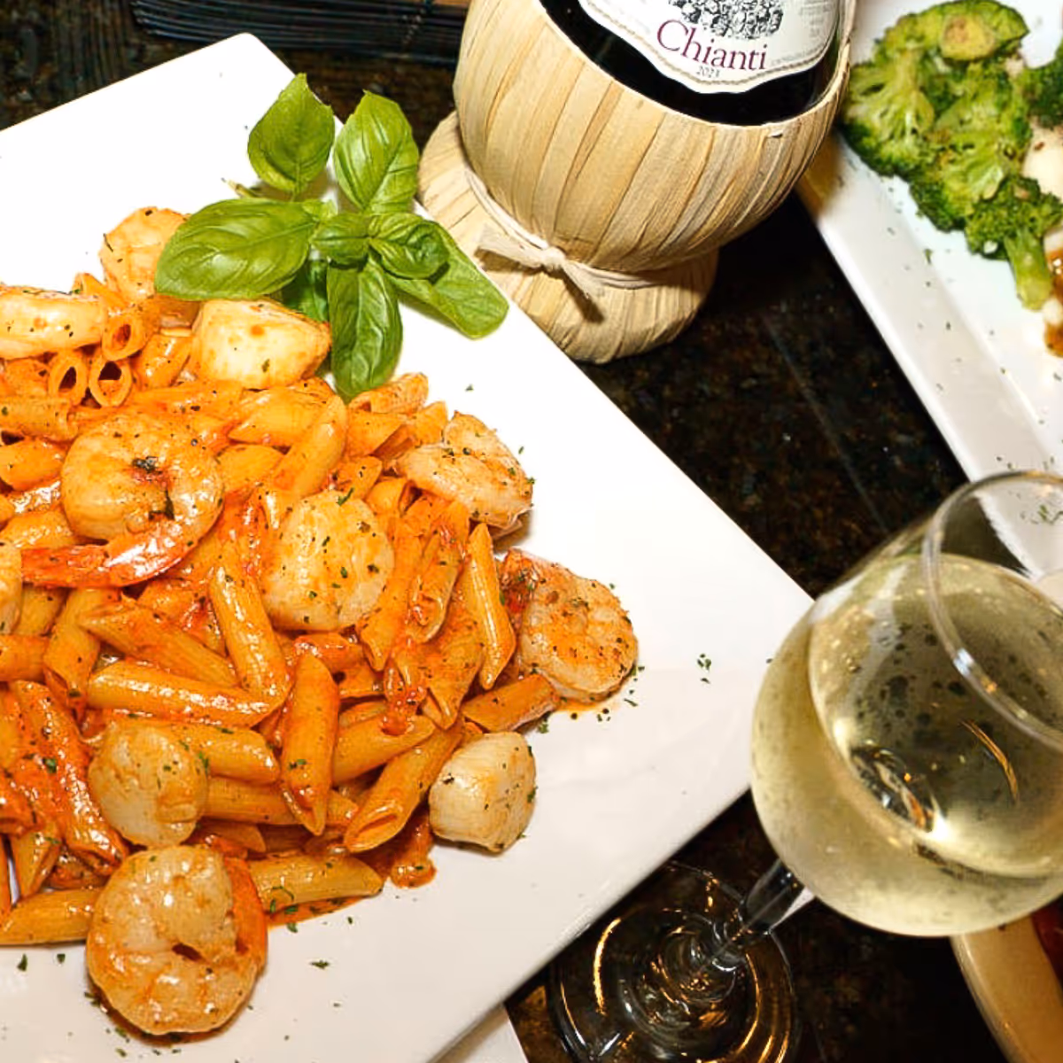 Plate of shrimp and penne pasta garnished with basil leaves, accompanied by a glass of white wine and a straw-covered bottle of Chianti wine.