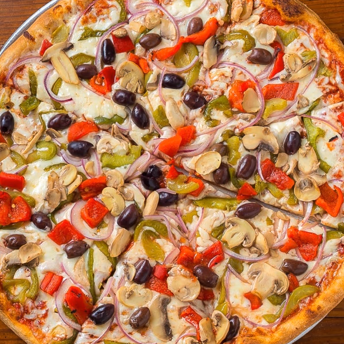 Pizza topped with black olives, red and green bell peppers, mushrooms, red onion, and melted cheese on a wooden table.