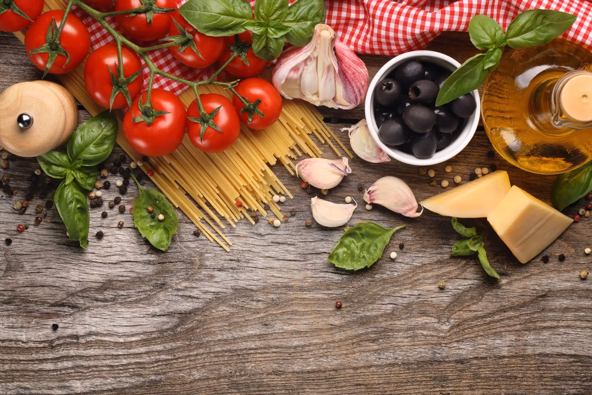 Cherry tomatoes on vine, uncooked spaghetti, garlic cloves, black olives in bowl, olive oil bottle, cheese wedges, basil leaves, and peppercorns on wooden surface with red checkered cloth.
