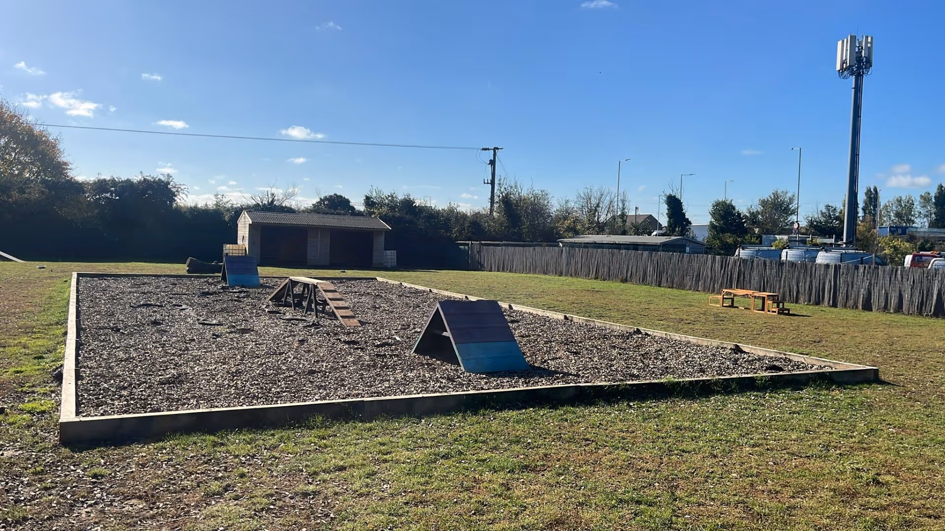 Outdoor dog agility training course with ramps, obstacles, and a wooden shed under a clear blue sky.