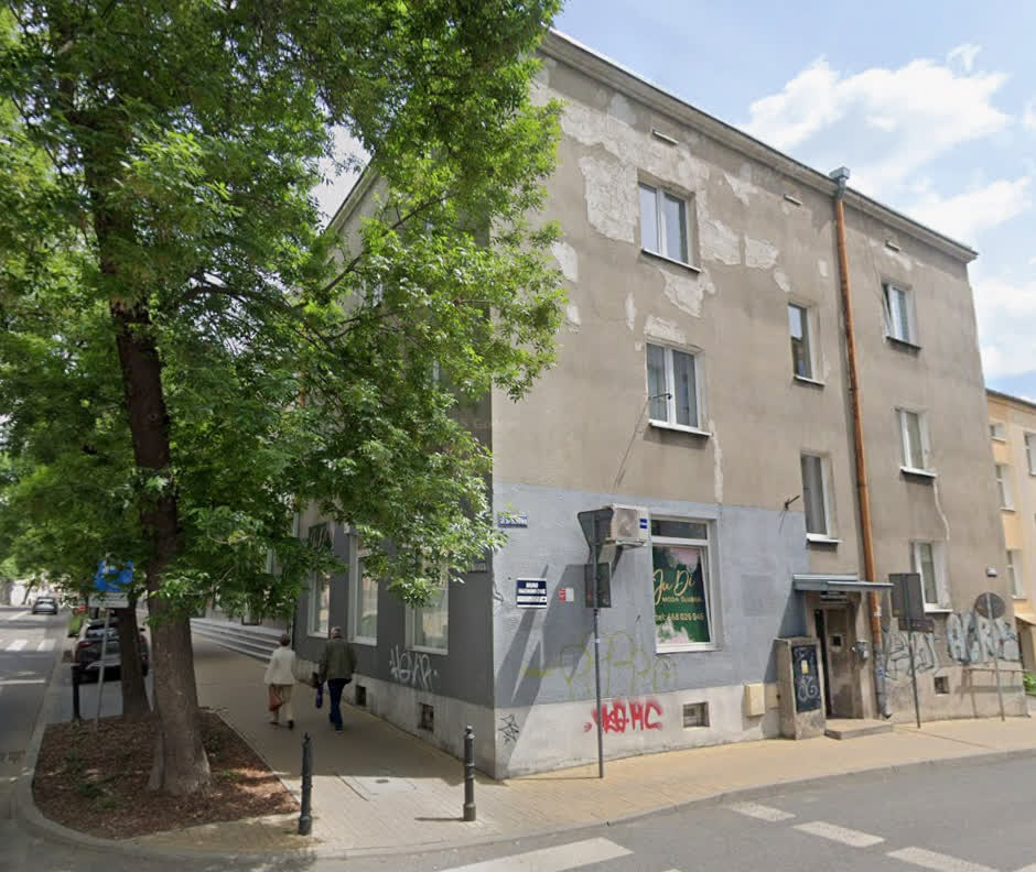 Three-story corner building with gray peeling facade, graffiti, and a large tree on the sidewalk with two people walking nearby.