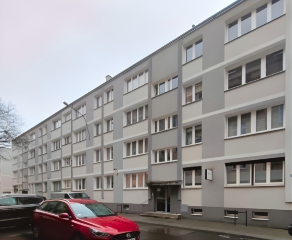 Street view of a modern five-story residential building with white and gray facade, parked cars, and a cloudy sky.