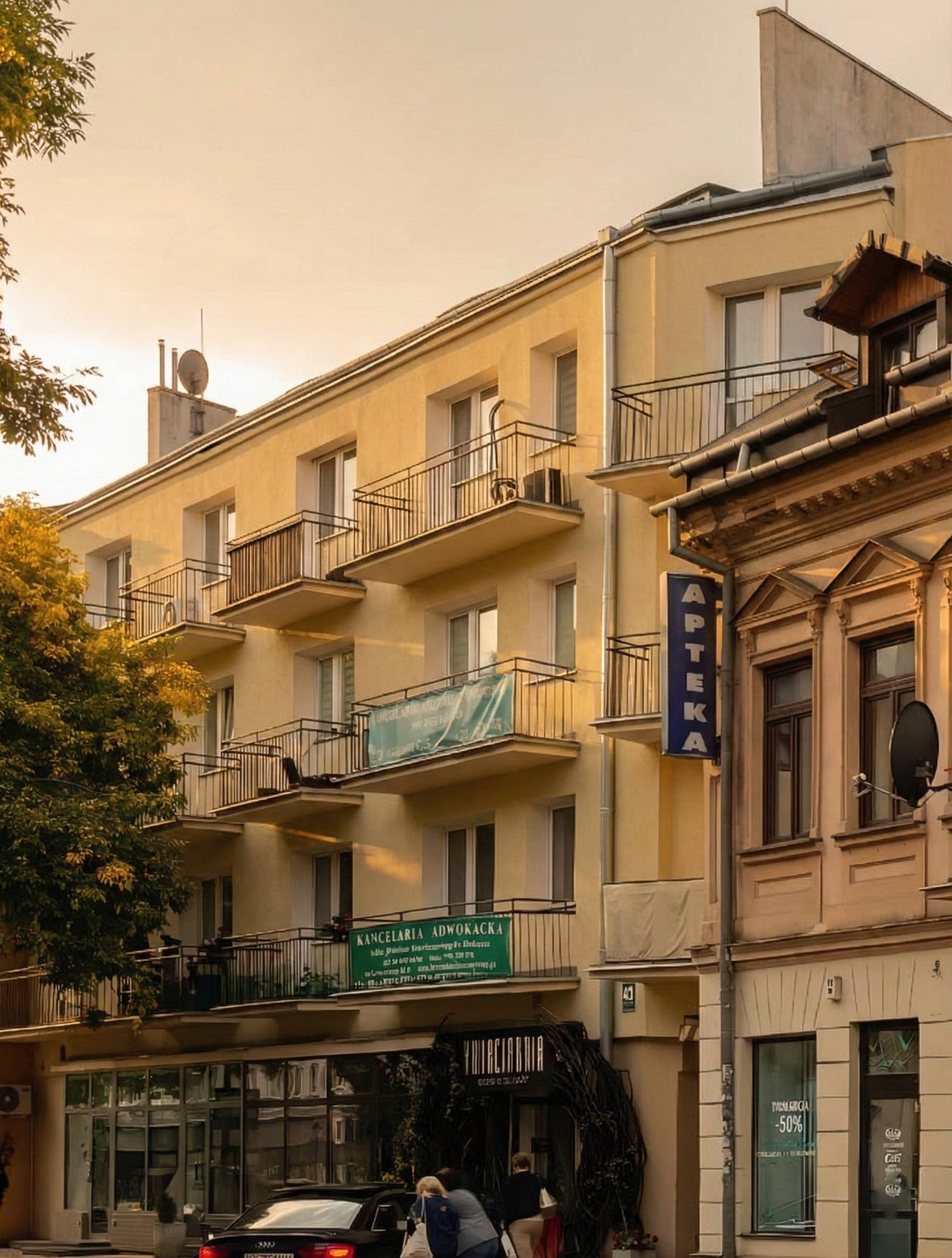 Yellow apartment building with balconies and stores below, including a pharmacy sign reading 'APTEKA', with people walking near a black car.