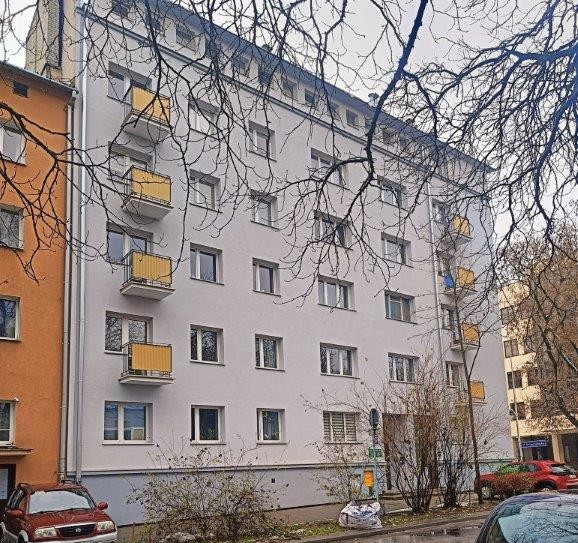 Five-story residential building with white facade and yellow balconies next to an orange building, with leafless tree branches overhead.