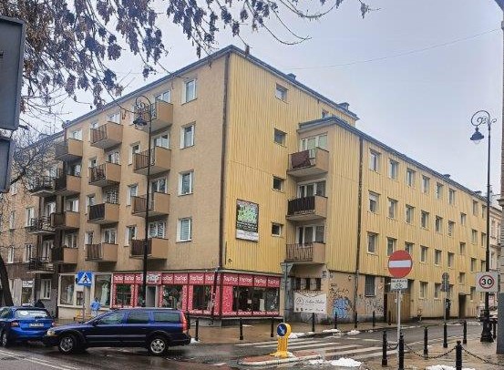 Four-story beige and yellow residential building with balconies on a street corner, cars parked nearby, and a no-entry traffic sign.