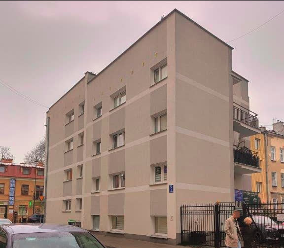 A four-story beige apartment building on a street corner with parked cars and two people walking nearby.