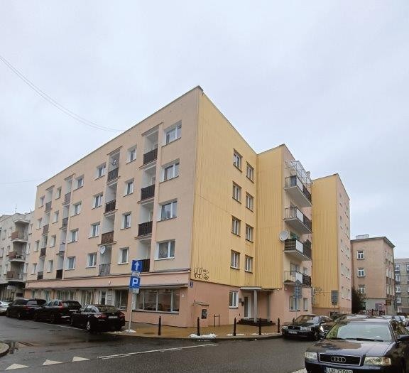 Four-story beige and yellow apartment building on a street corner with parked cars and cloudy sky.
