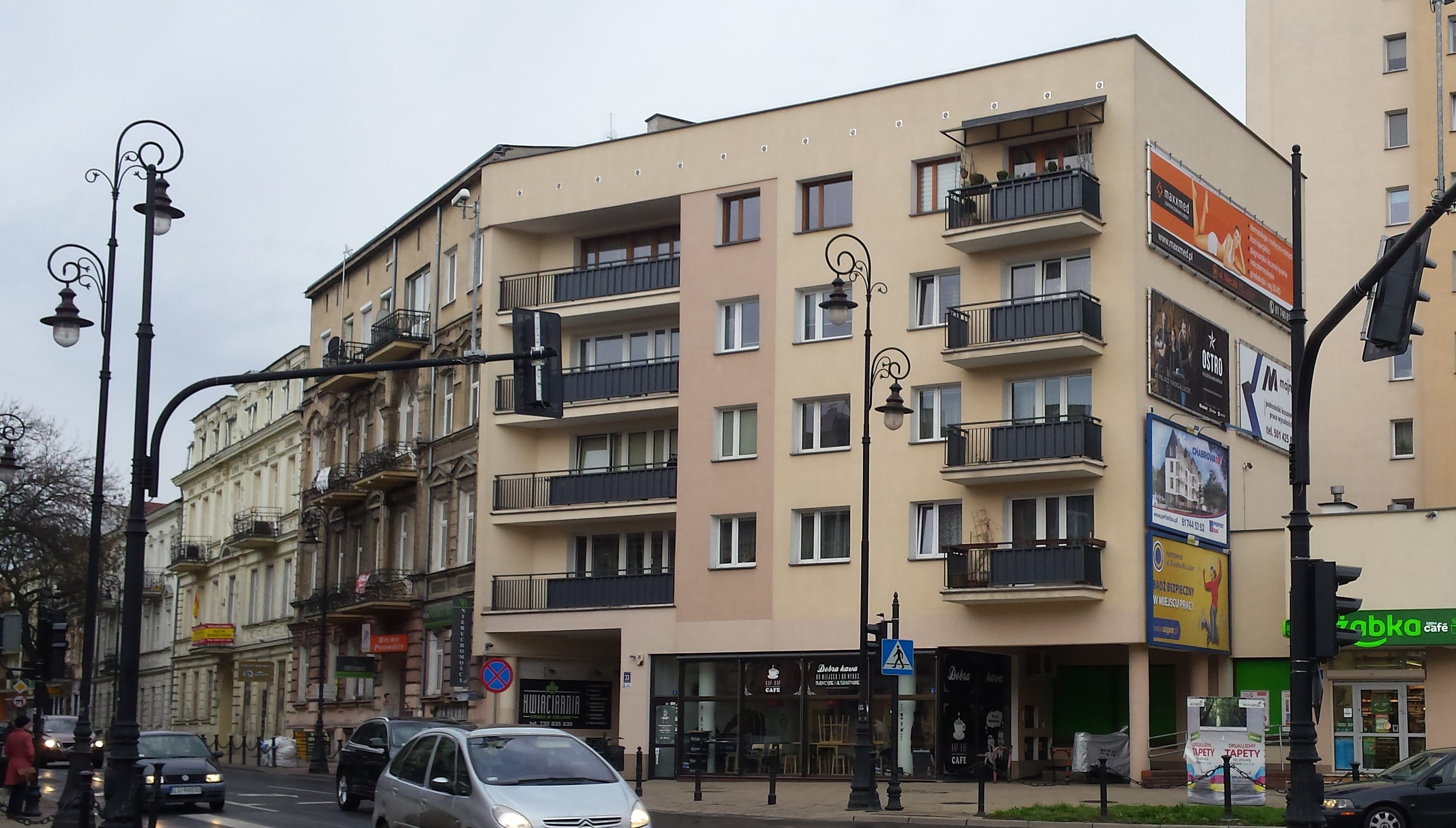 Street view of a corner building with balconies, street lamps, cars, and various shop signs including a café and Żabka store.
